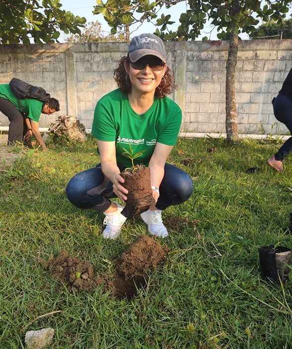 Mujer con camisa verde plantando un retoño en una zona de césped al aire libre, sonriendo y con gorra y guantes.