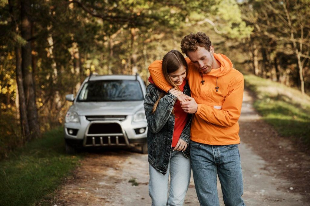 Una pareja camina por un camino de tierra junto a un SUV blanco en una zona boscosa.