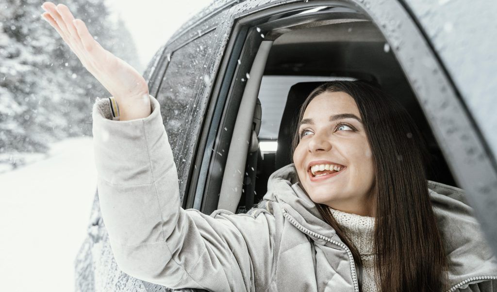 Mujer sonriendo y saludando desde la ventanilla de un coche en un día nevado.