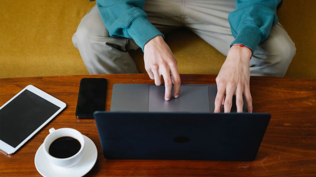 Una persona usa una computadora portátil en una mesa de madera con una tableta y una taza de café cerca.