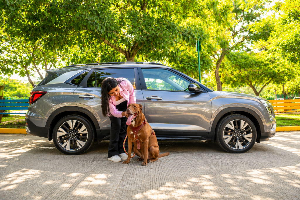 Mujer con dos perros de pie junto a un SUV gris en una carretera arbolada.