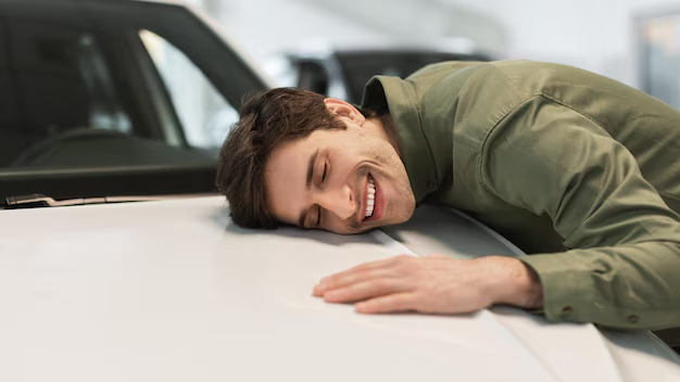 Hombre sonriendo con la mejilla apoyada en el capó blanco de un coche en una sala de exposición.