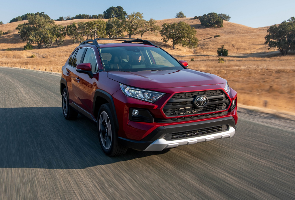 Un SUV rojo circula por una carretera rural a través de colinas áridas bajo un cielo despejado.