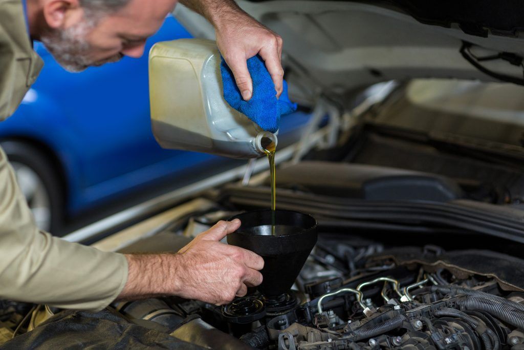 Un mecánico vierte aceite en el motor de un coche con un recipiente azul debajo del capó abierto.