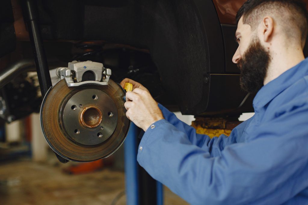 Mecánico con chaqueta azul inspeccionando un disco de freno de coche con una herramienta en un taller mecánico.