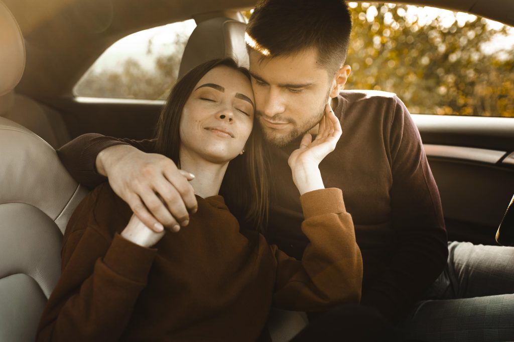 Una pareja se abraza en el asiento trasero de un coche; la mujer sonríe y descansa en los brazos del hombre.