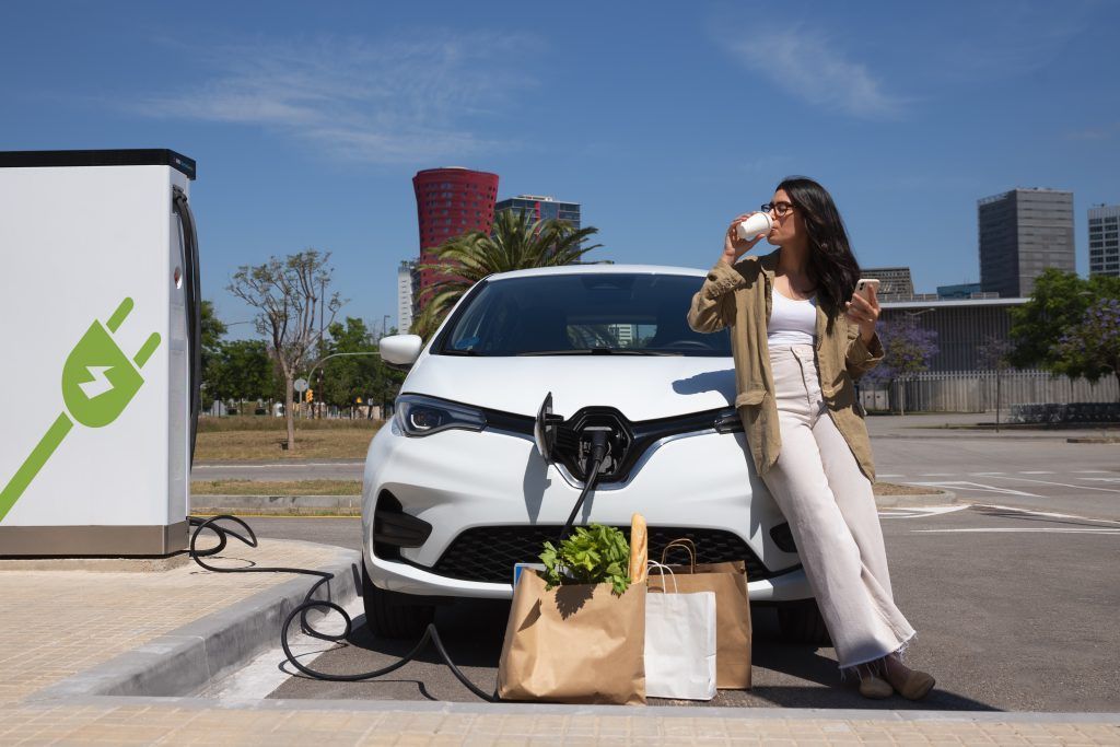 Mujer cargando un vehículo eléctrico blanco al aire libre junto a una estación de carga y bolsas de papel para la compra.