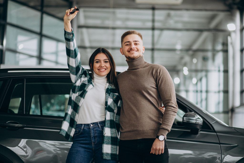 Dos personas de pie junto a un coche, sonriendo y levantando las llaves en una luminosa sala de exposición interior.