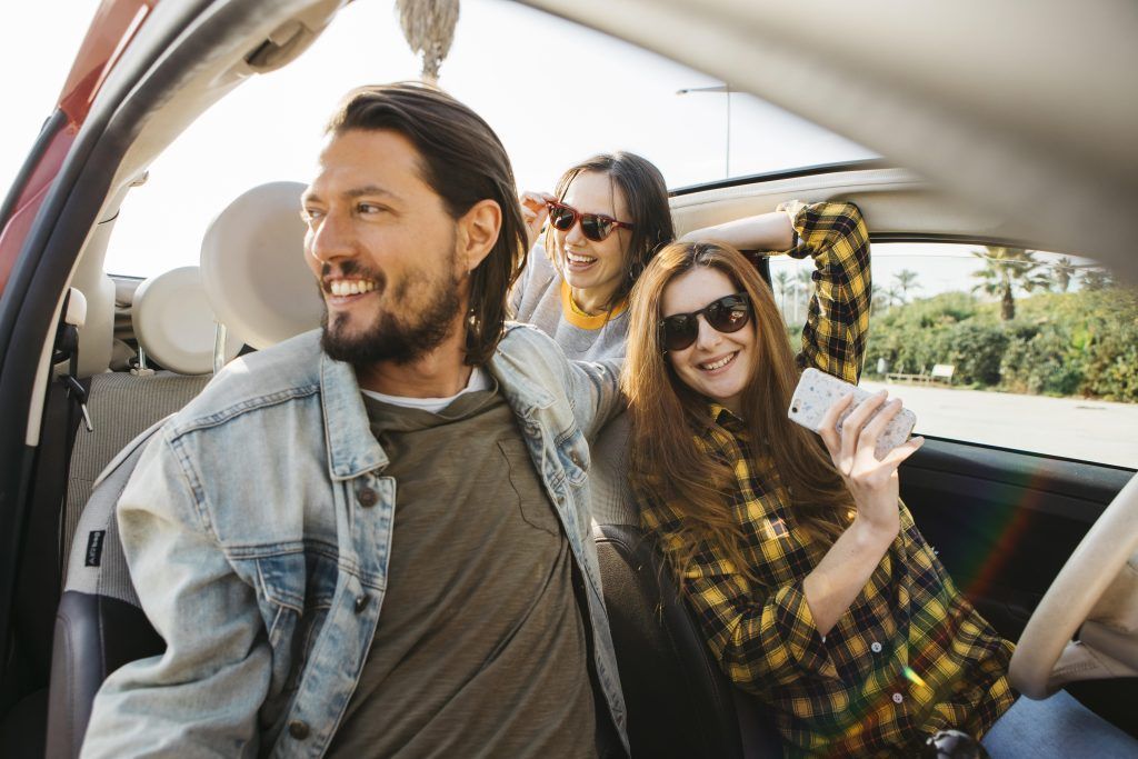 Tres amigos sonriendo en un coche, uno de ellos asomado por la ventanilla abierta con gafas de sol.