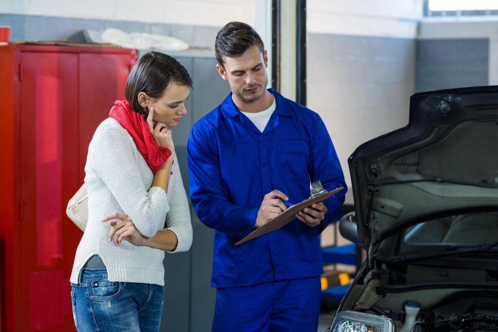 Un mecánico le muestra un portapapeles a un cliente junto a un coche con el capó abierto en un garaje.