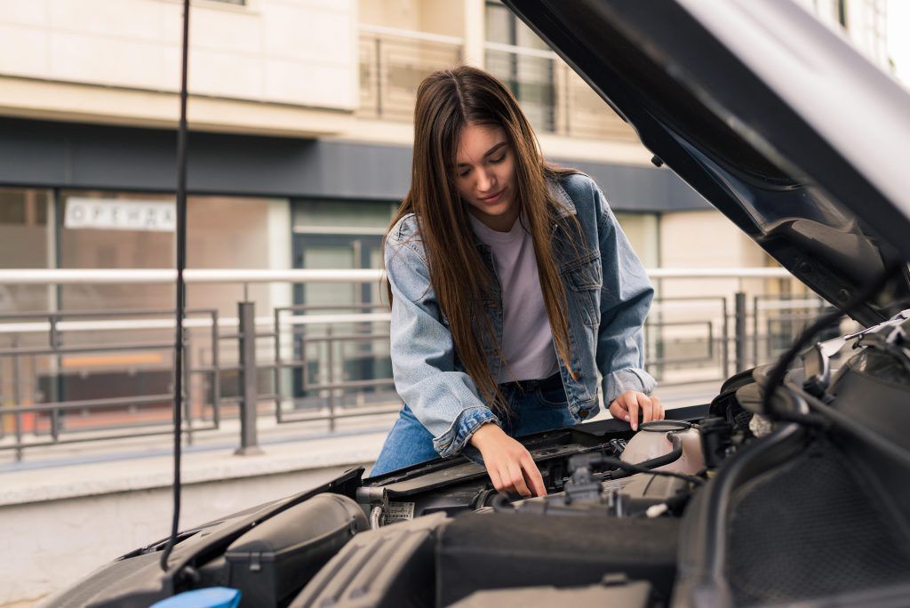 Mujer inspeccionando el motor de un coche bajo el capó abierto en un aparcamiento.