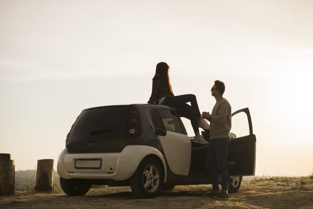 Dos personas junto a la puerta abierta de un coche al atardecer, una de ellas sentada en el techo del vehículo.