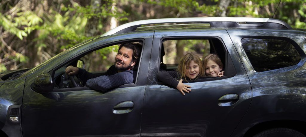 Familia sonriendo y saludando desde una camioneta oscura que circula por una zona boscosa.