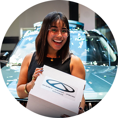 Mujer sonriendo y sosteniendo una caja frente a un coche en una exposición interior.