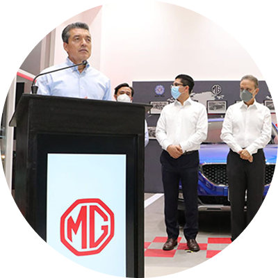 Un hombre habla desde un podio de MG, con dos empleados enmascarados de pie junto a una exposición de coches.