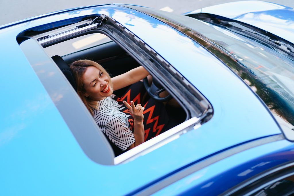 Mujer sonriente recostada dentro de un auto azul vista a través del techo corredizo.
