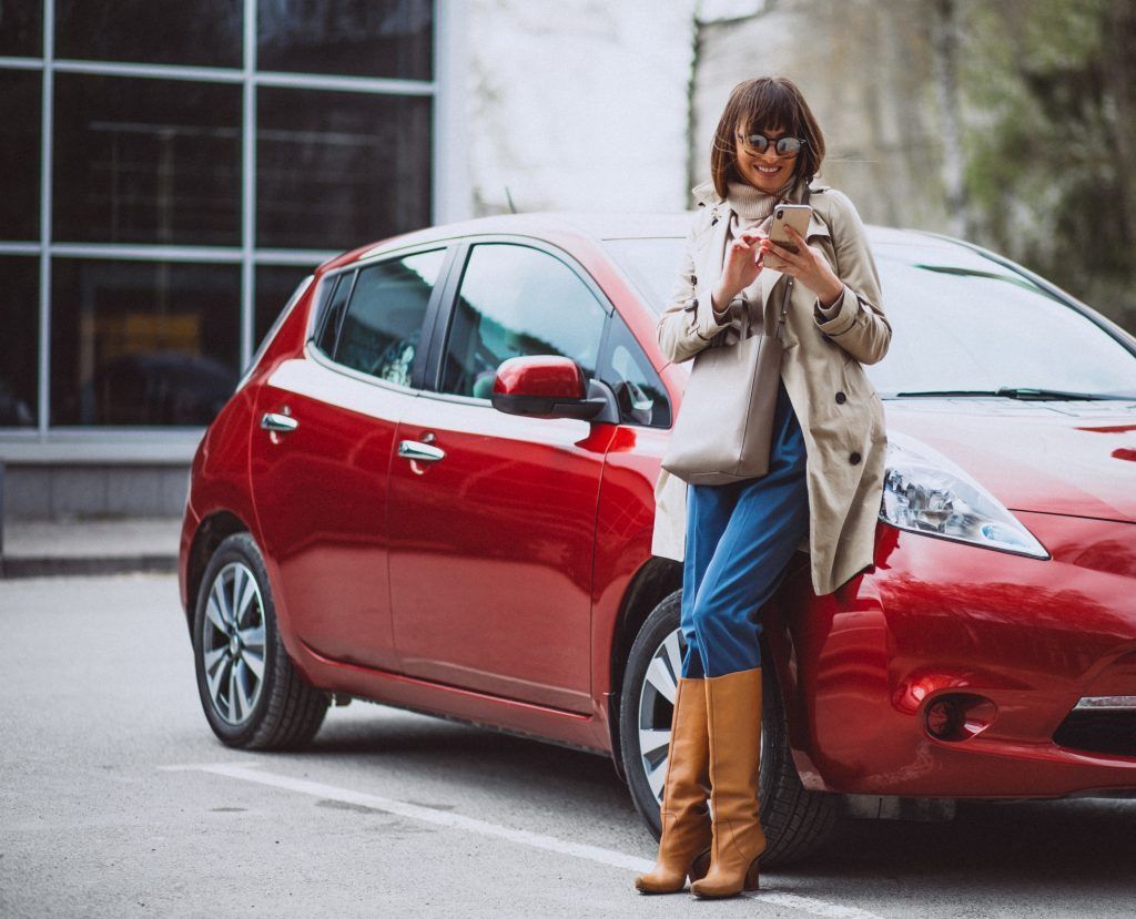 Mujer de pie junto a un coche rojo, mirando su teléfono en una calle de la ciudad.
