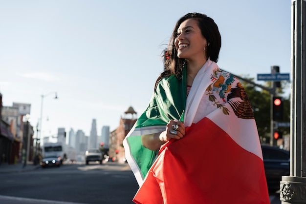Una persona envuelta en una bandera mexicana sonríe en una calle de la ciudad al atardecer.