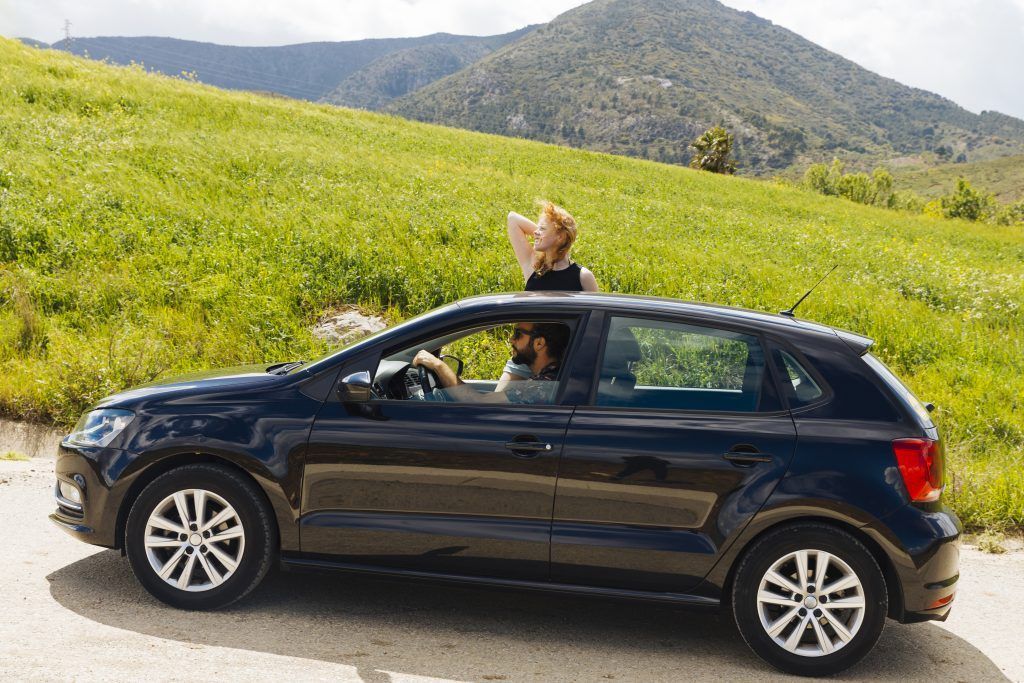 Un coche compacto negro en una carretera rural, con un perro asomado por el techo solar y colinas verdes de fondo.