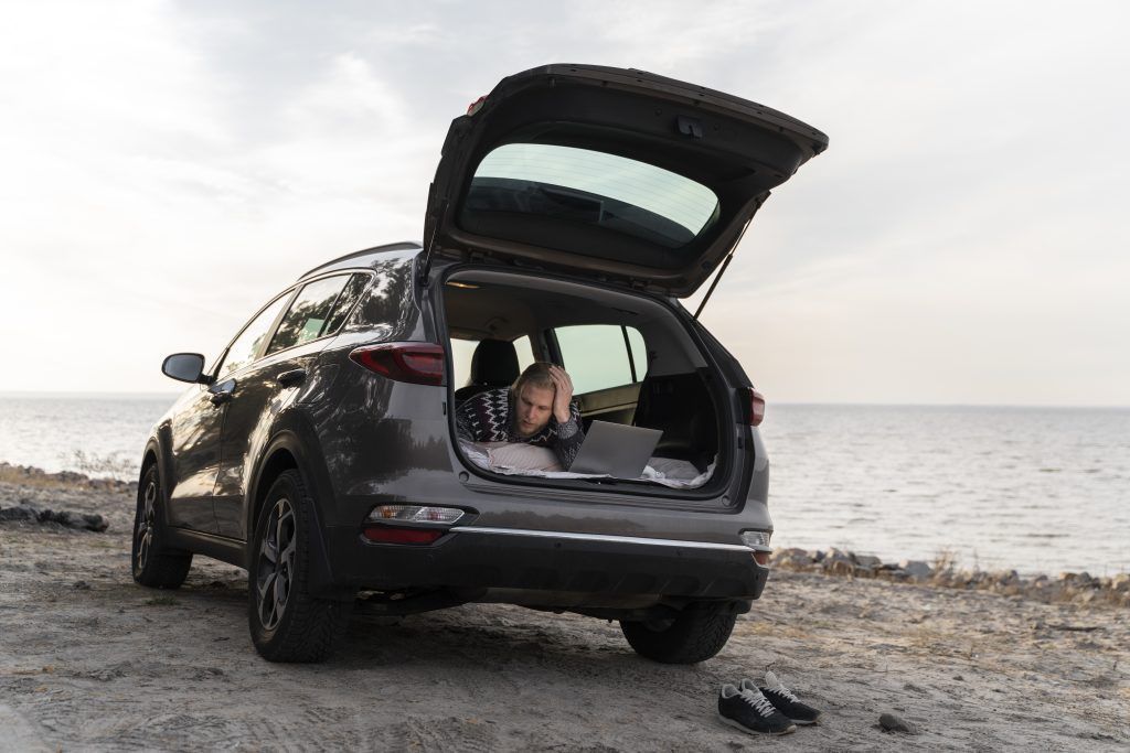 Un auto con el portón trasero abierto en una playa rocosa al atardecer, con una persona de pie junto al maletero abierto.