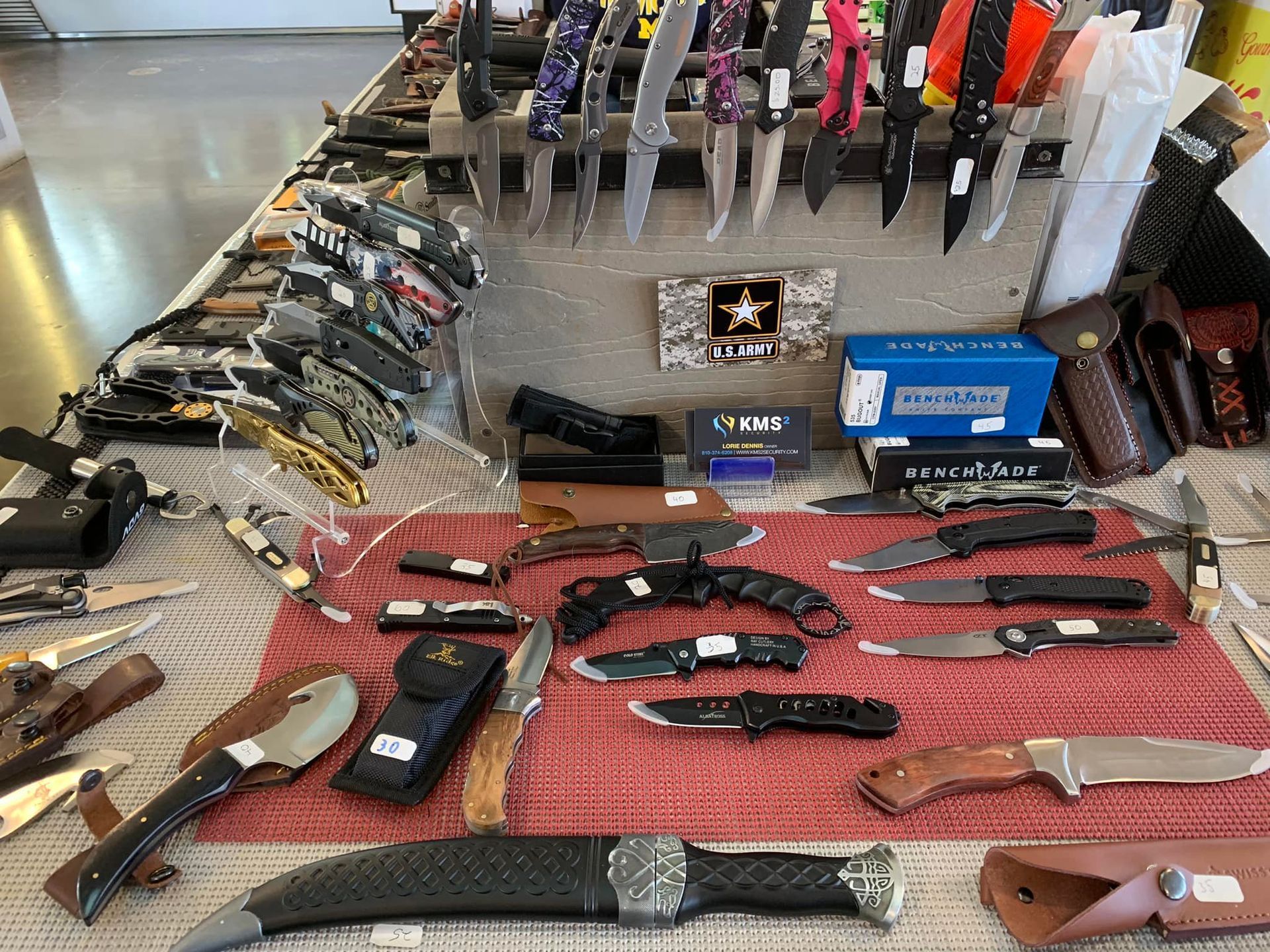 Display of various knives on a table at a sale. Includes folding knives, fixed-blade knives, and sheaths.