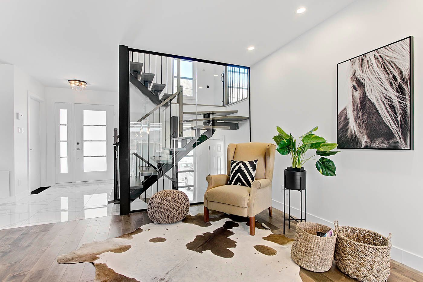 Entrée moderne et lumineuse avec un escalier en verre, un fauteuil, un tapis en peau de vache, des plantes et des paniers tressés