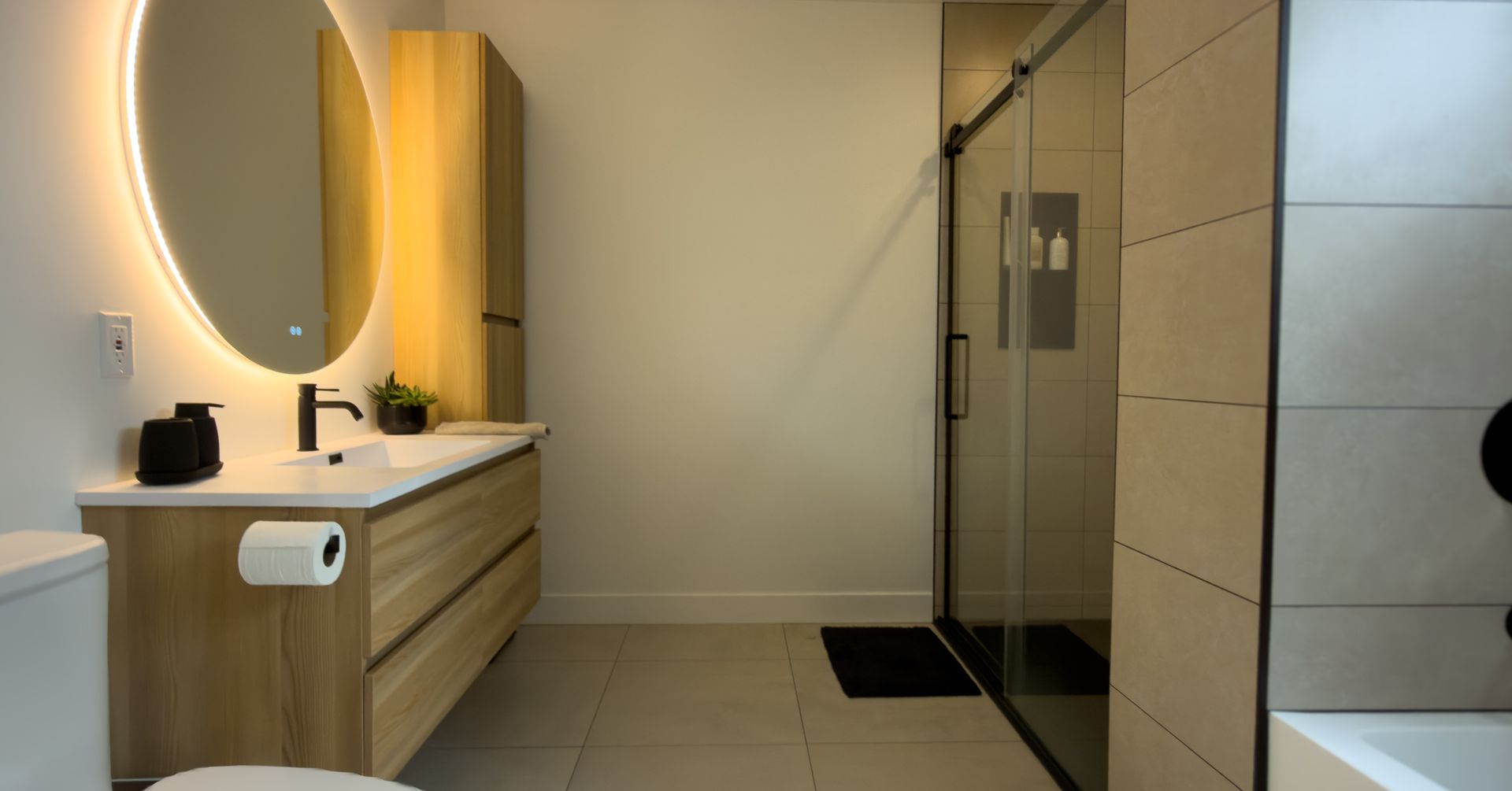 Salle de bains moderne avec meuble-lavabo suspendu en bois, miroir ovale éclairé, douche vitrée et baignoire.