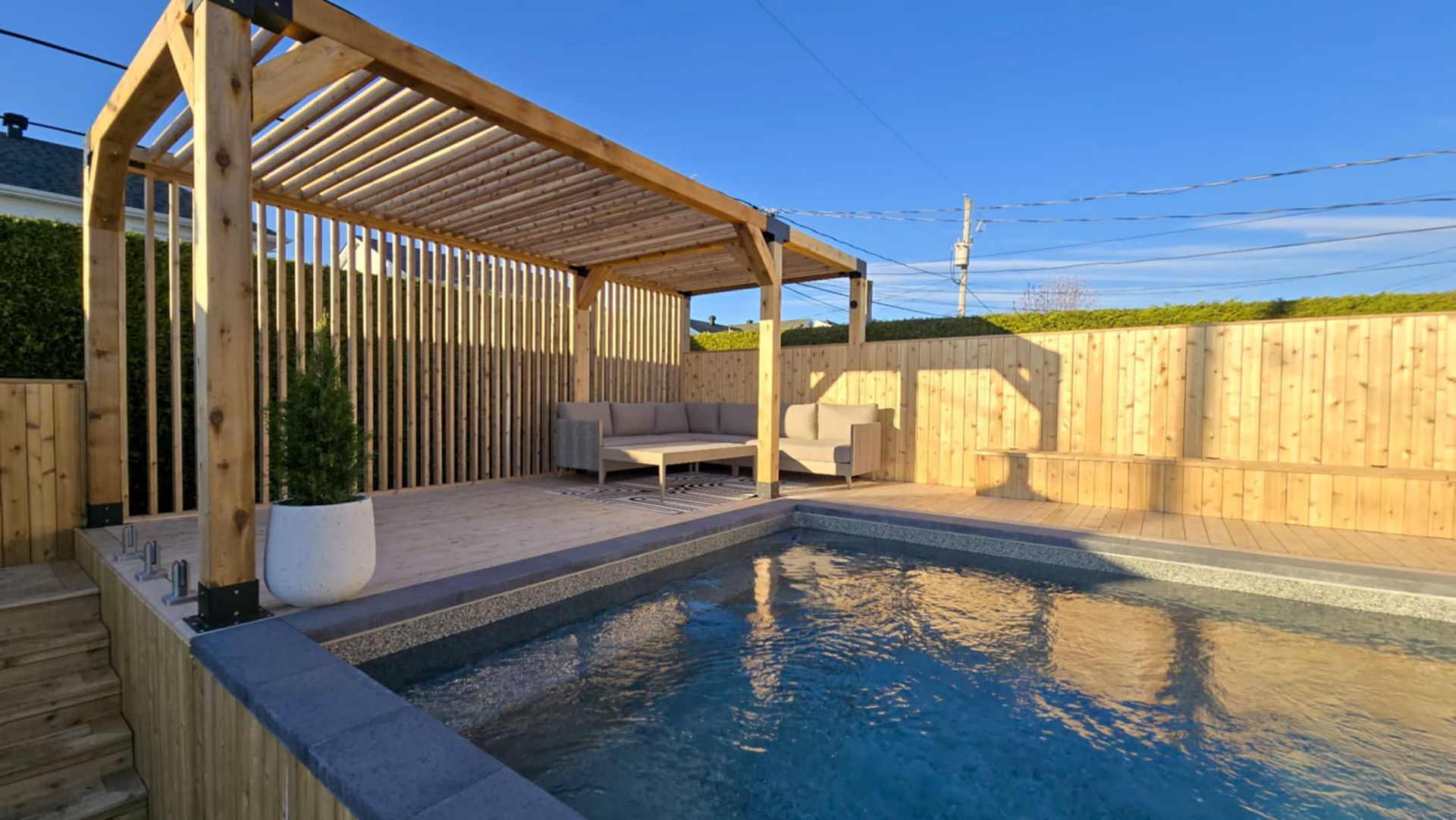 Piscine dans un jardin avec pergola en bois, chaises longues et patio clôturé baigné d’un doux soleil