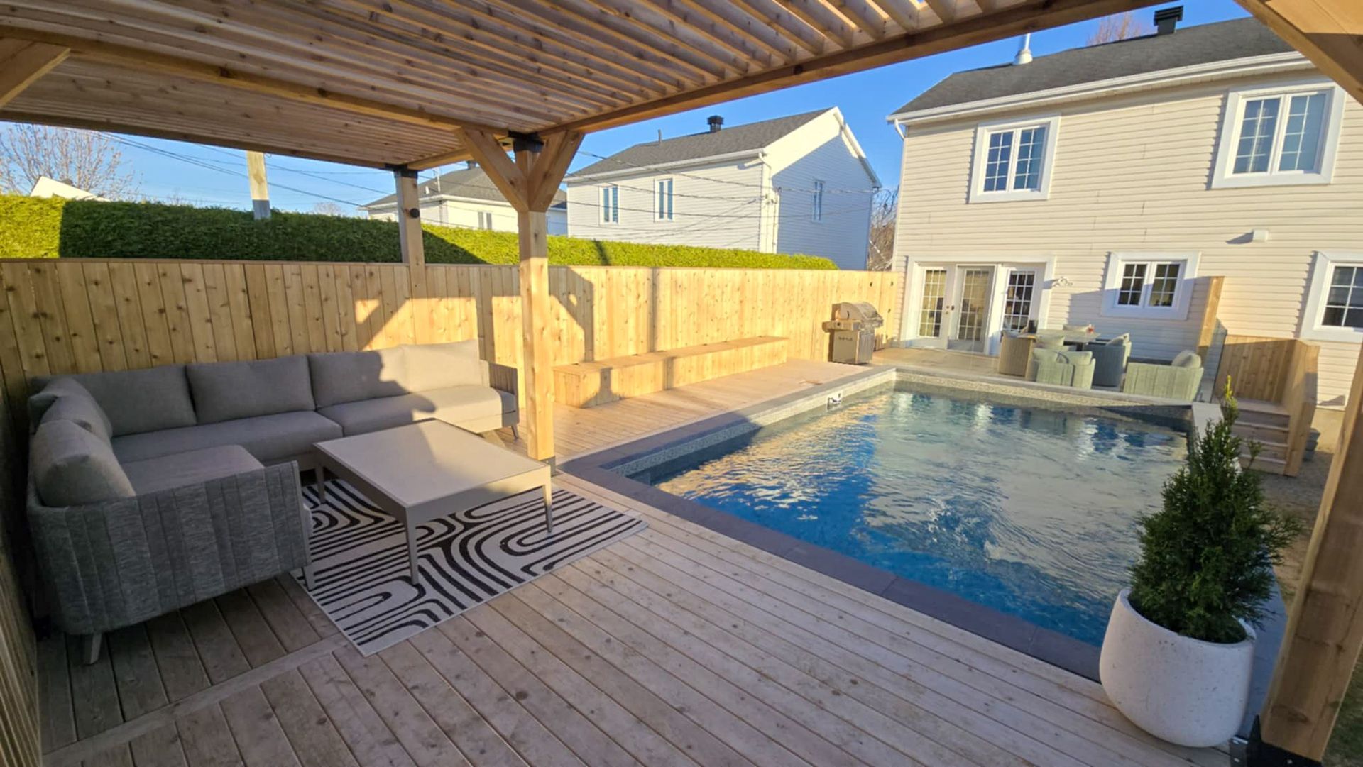 Patio couvert avec un salon de jardin gris donnant sur une piscine dans le jardin, à côté d’une maison beige.