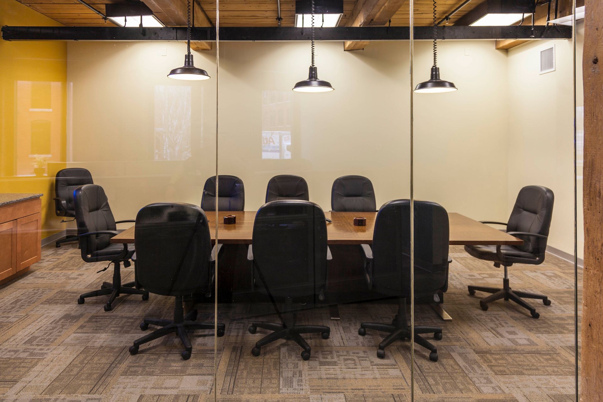 A modern, empty glass-walled conference room featuring a large wooden table and black swivel chairs under pendant lights.