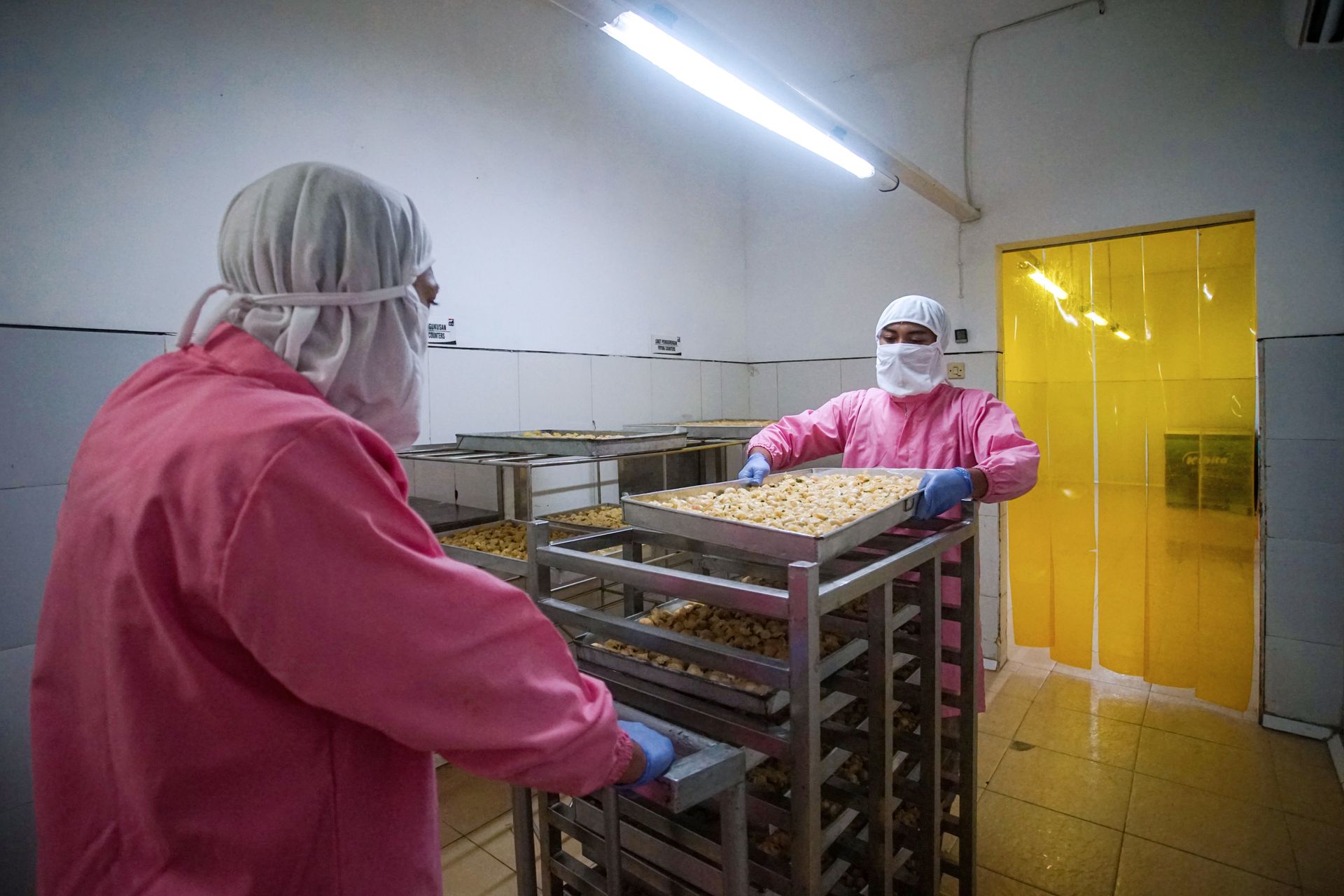 Two workers in pink protective gear and face masks move a metal tray cart filled with food inside a brightly lit facility.