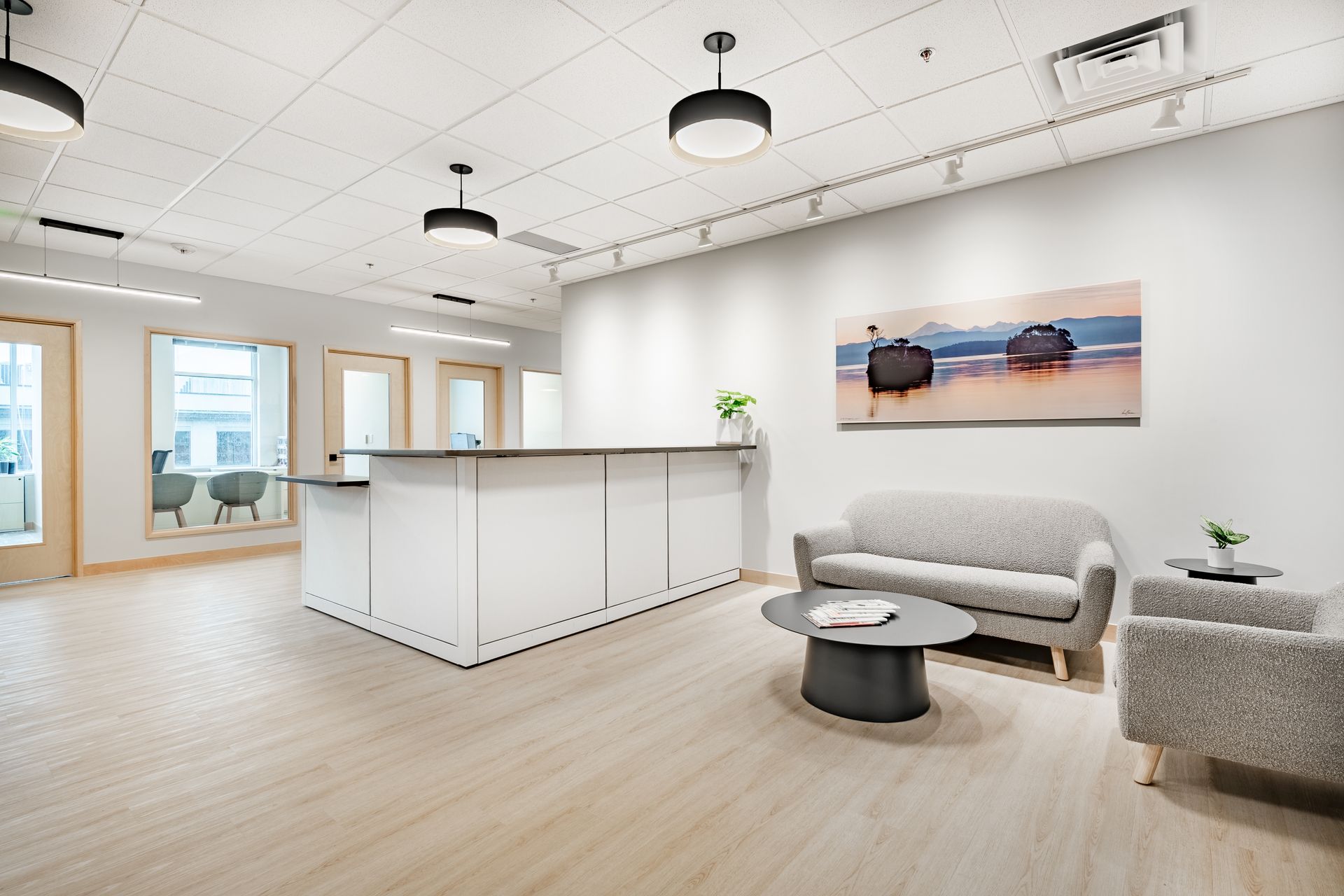 A modern, bright office lobby featuring a white reception desk, gray sofa and armchairs, and a beach painting on the wall.