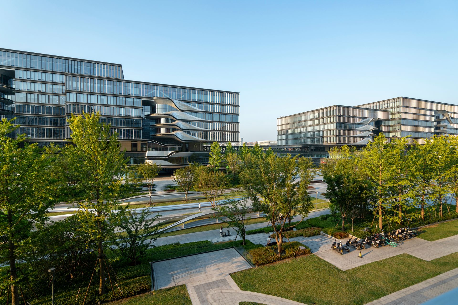Modern office buildings with glass facades overlooking a lush, landscaped park with trees and stone paths.