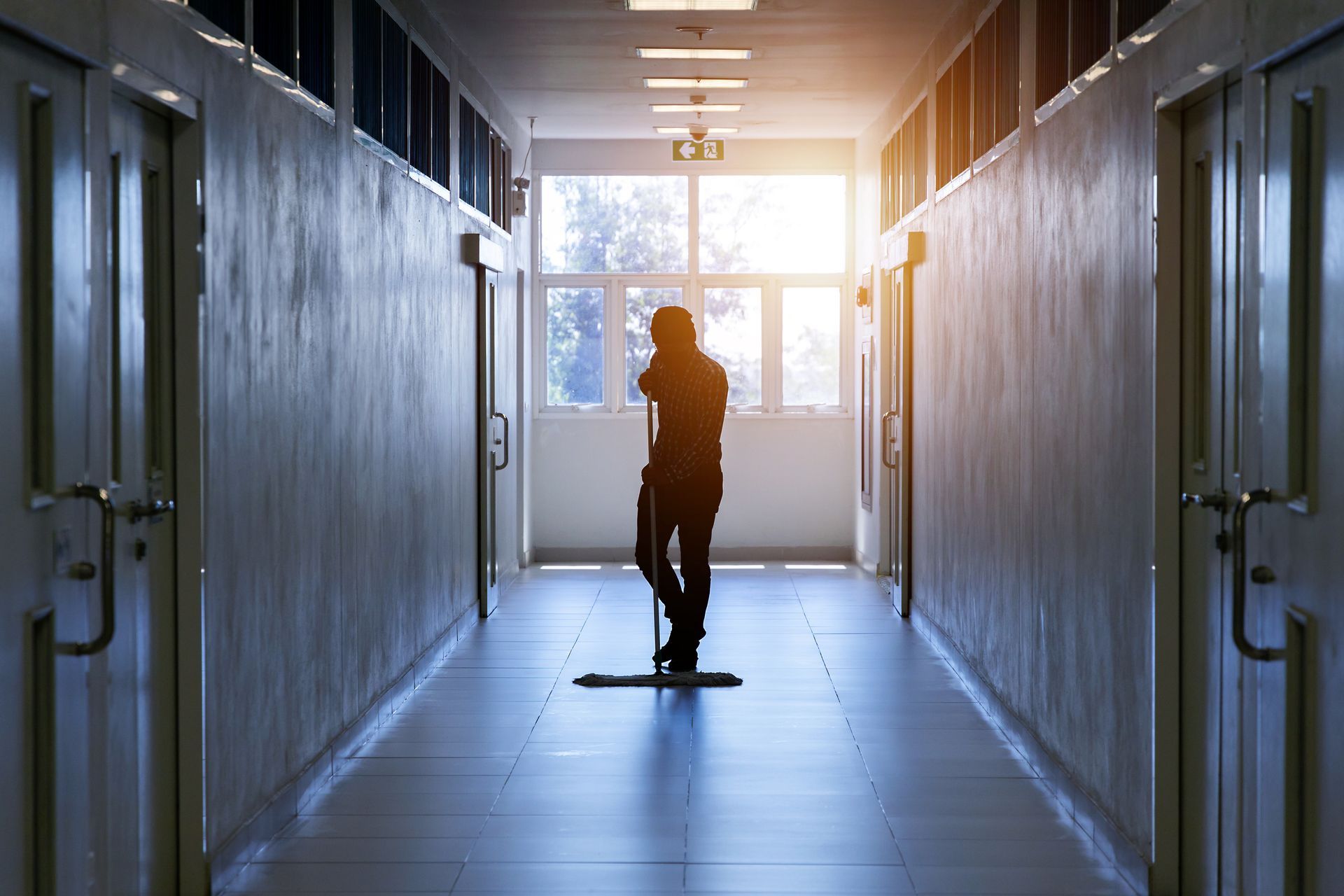 A silhouette of a person mopping a long, sunlit hallway lined with doors.