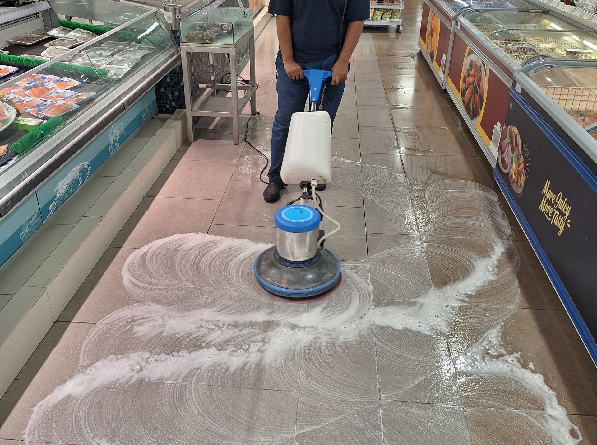 A person using a floor scrubbing machine to clean a grocery store aisle covered in white soapy cleaning solution.