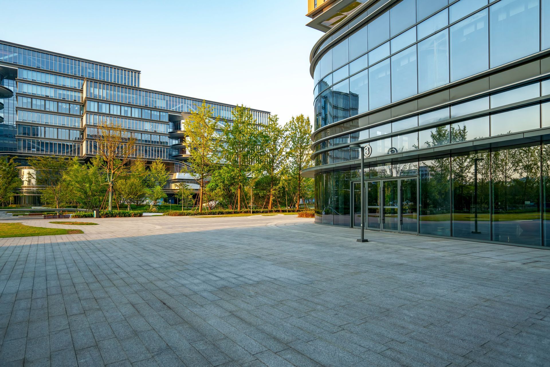 Modern glass buildings with a stone plaza and trees.