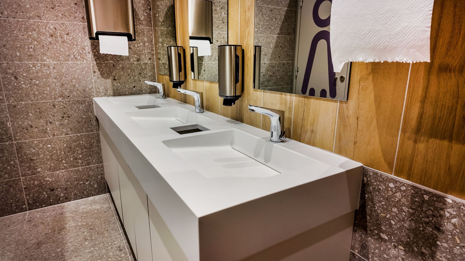 Public restroom with a long white sink, silver faucets, and dispensers. Gray and wood-tone walls.