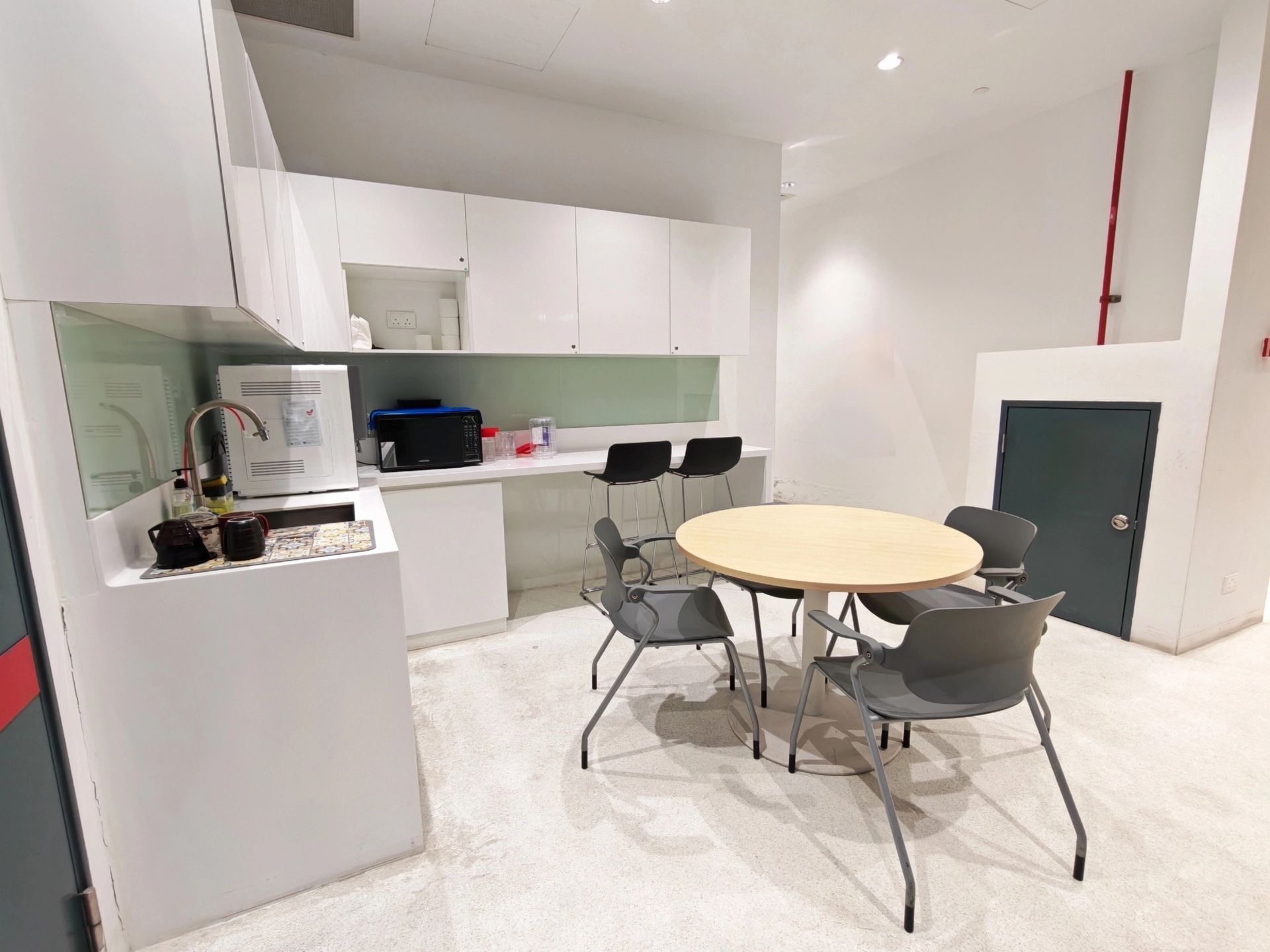 A small break room with white cabinets, a sink, a round table, and four gray chairs on a light-colored floor.