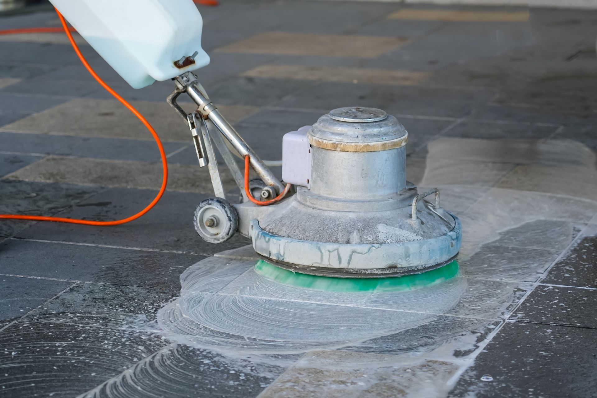 A floor buffer machine with a cleaning solution tank scrubs a gray tiled floor, creating a soapy lather.