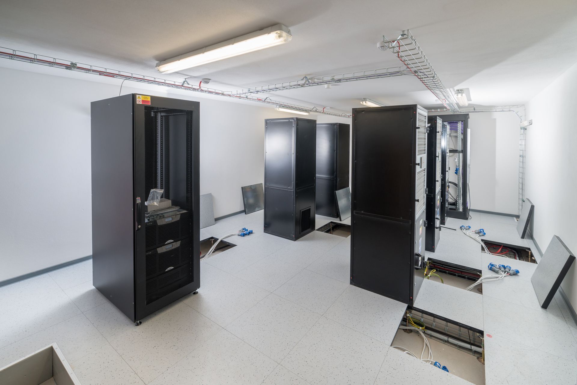Server room with several black equipment racks installed on a raised floor with some floor tiles removed.