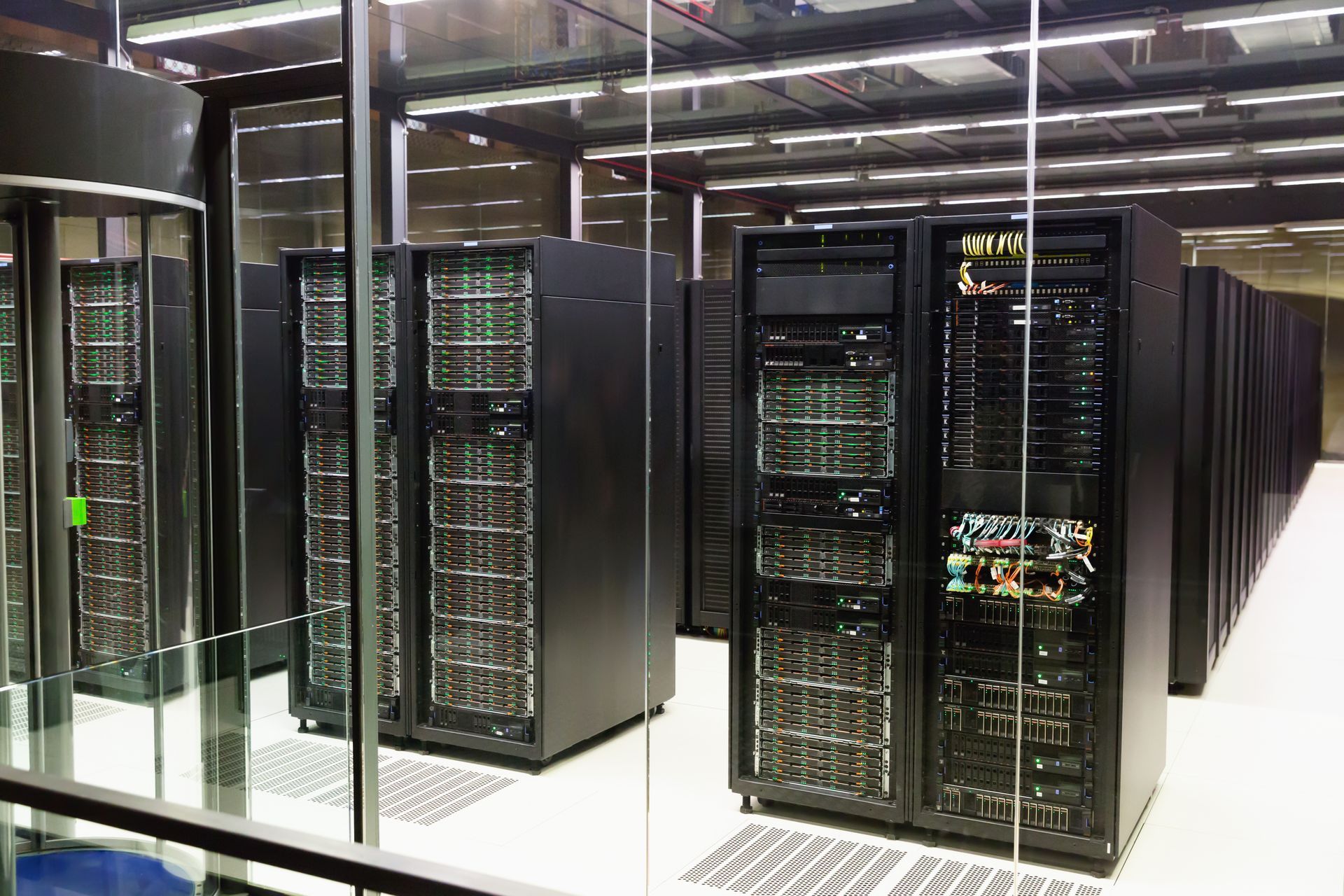 Rows of black server cabinets lined up in a brightly lit, modern data center, viewed through a glass partition.