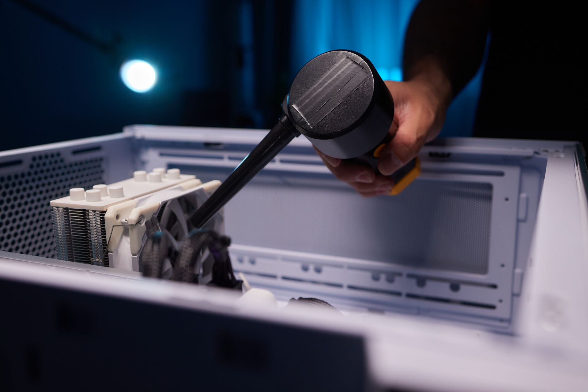 A person uses an electric air duster to clean dust from a computer CPU cooler inside a white PC case.