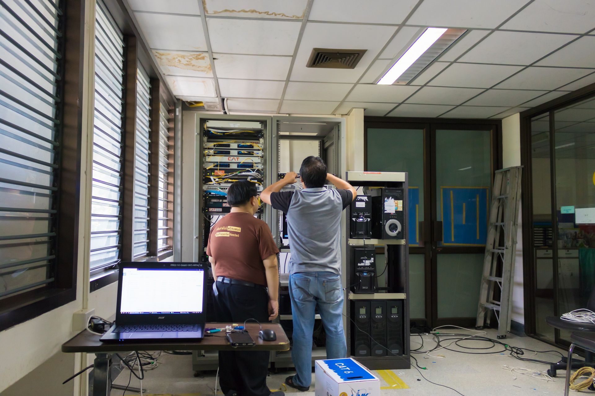 Two people work on IT equipment in server racks in an office room with a laptop on a table nearby.