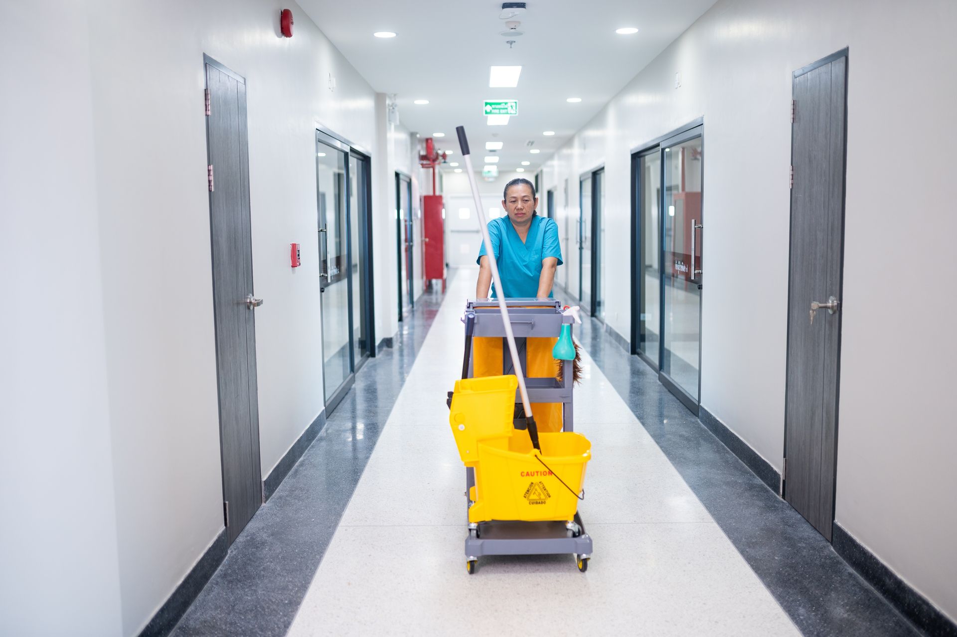 A custodian in scrubs pushes a yellow mop bucket and cleaning cart down a brightly lit hospital corridor.