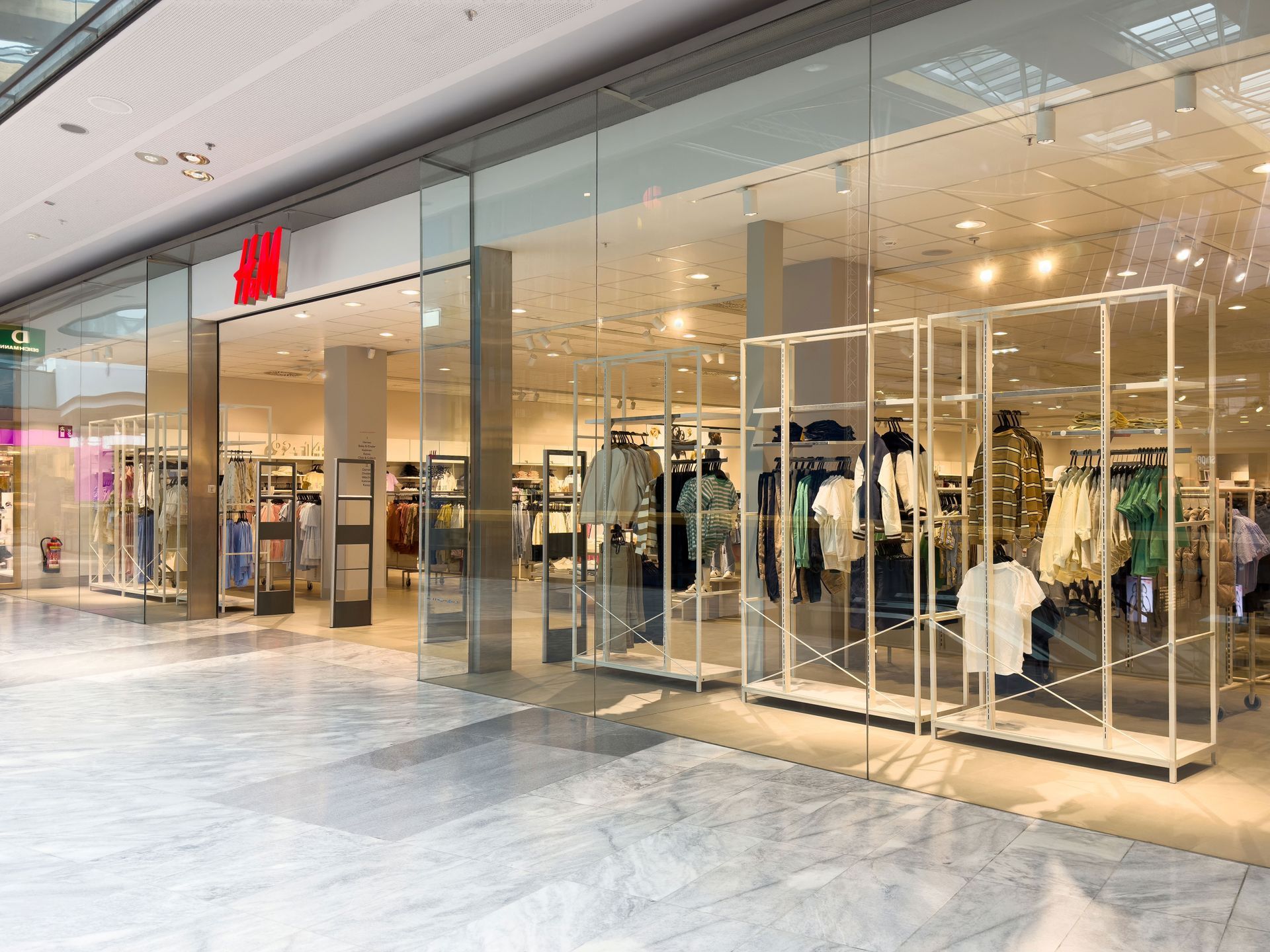 A wide-angle view of an H&M storefront inside a shopping mall with large glass windows and clothes on display racks.