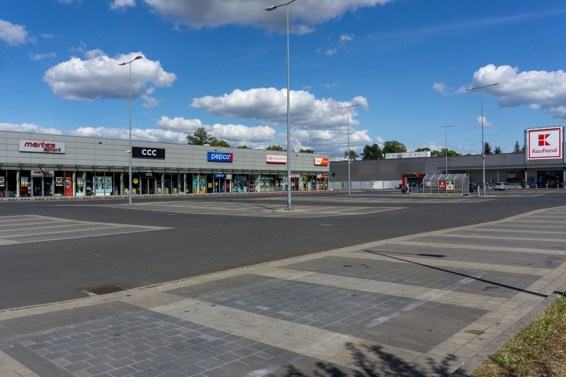 A retail strip mall with a large empty parking lot under a blue sky with clouds.