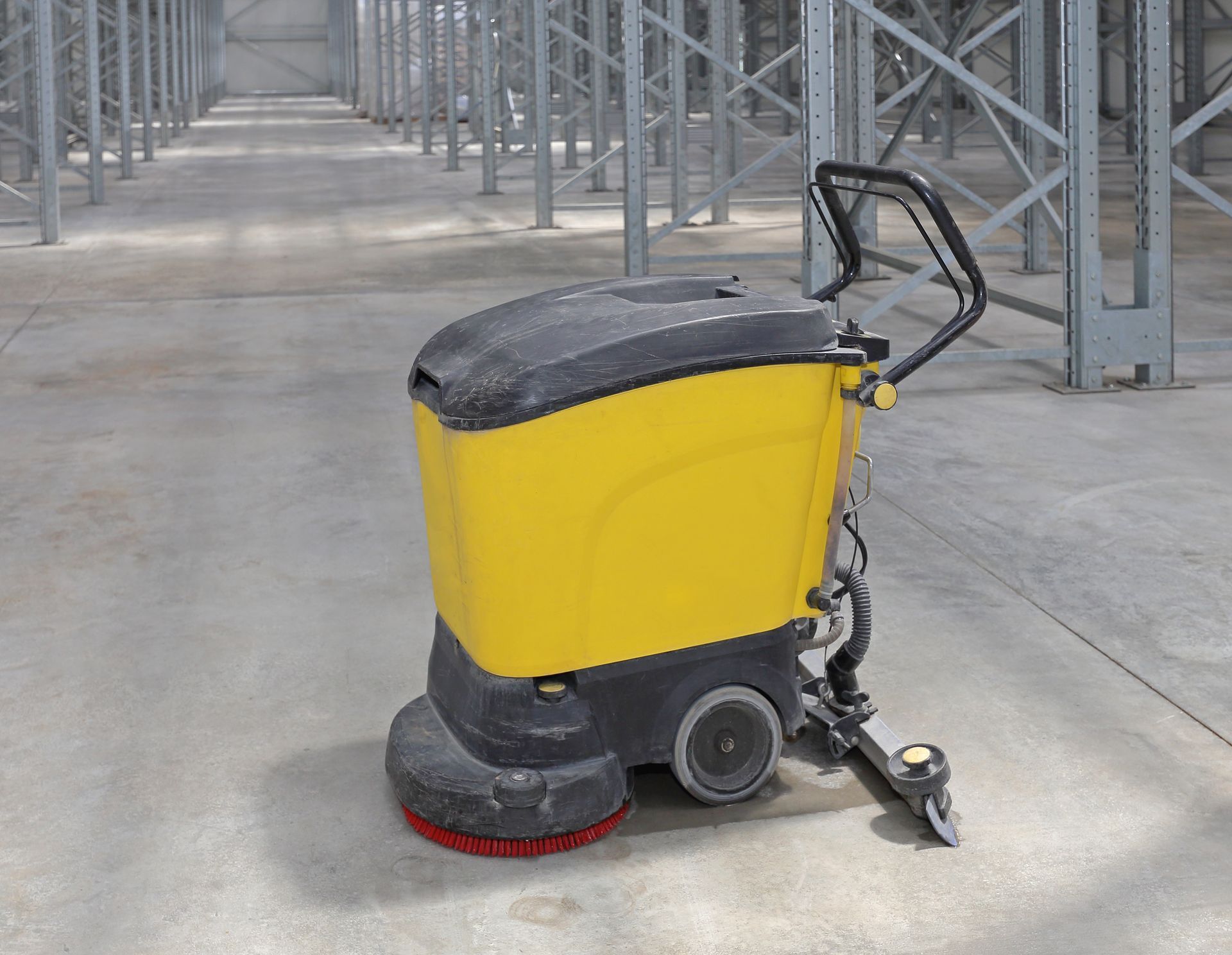 A yellow industrial floor scrubber parked on a concrete floor inside a warehouse with metal shelving.