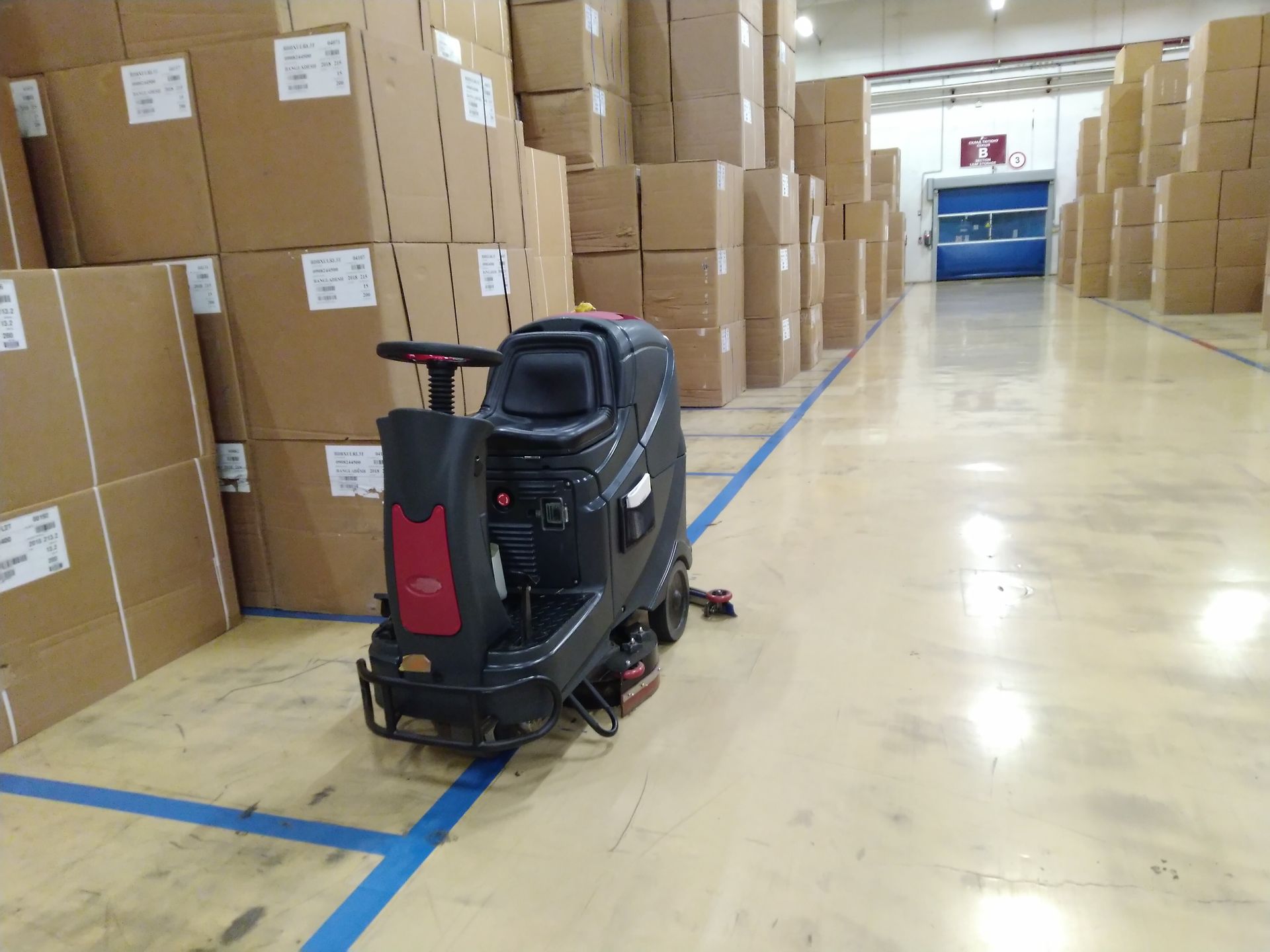 A floor scrubber machine parked in the aisle of a warehouse stacked with cardboard boxes.
