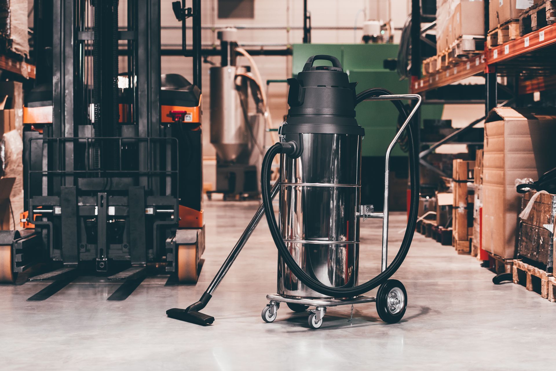 A tall, industrial metal vacuum cleaner stands on the floor of a warehouse next to a forklift and storage shelving.
