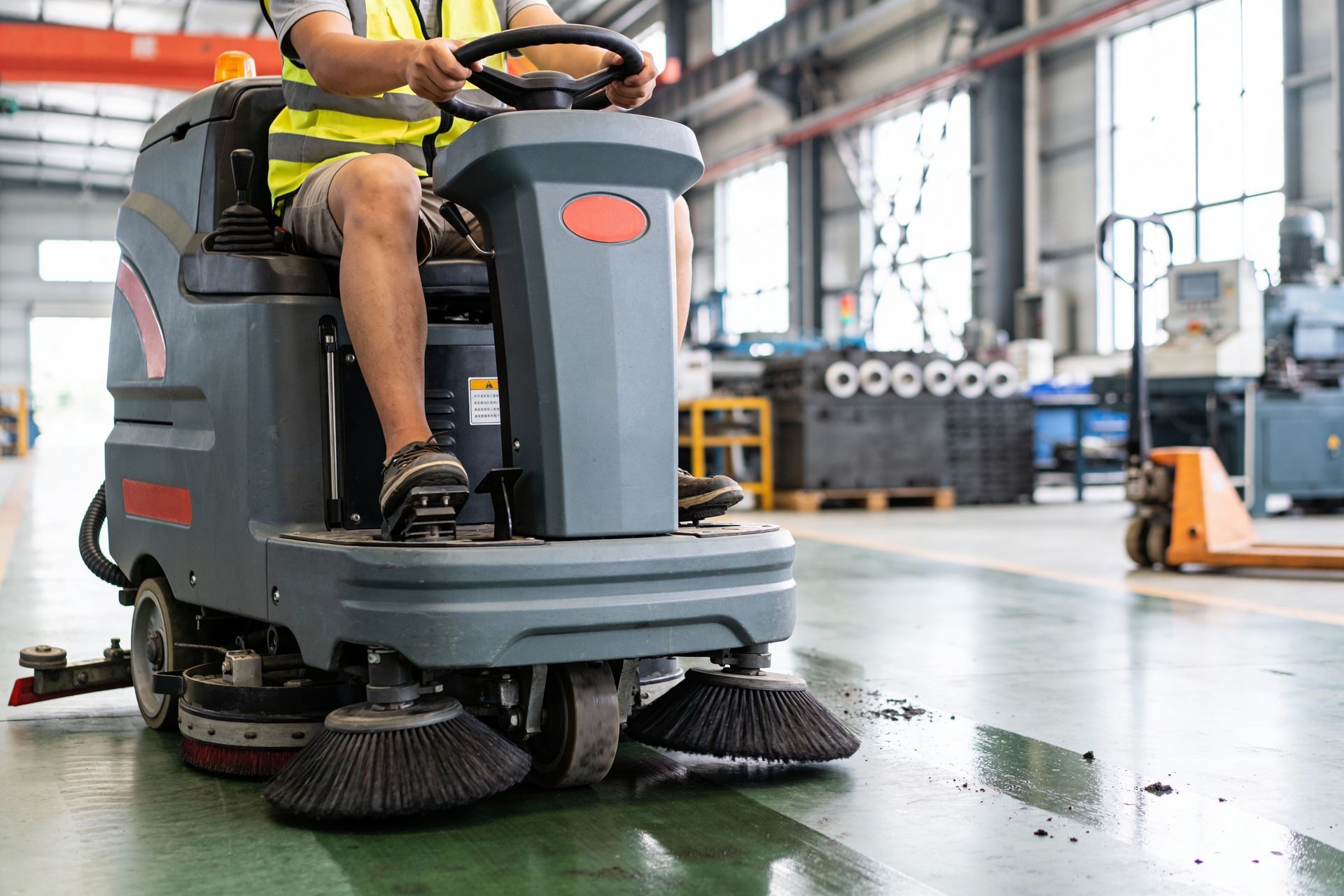 A person in a high-visibility vest operates a ride-on industrial floor scrubber in a warehouse.